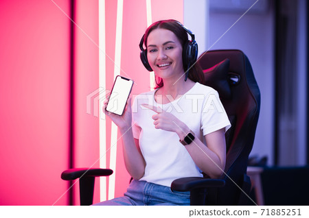 Cheerful gamer girl pointing with finger at smartphone with blank screen while sitting on a gaming chair. 71885251
