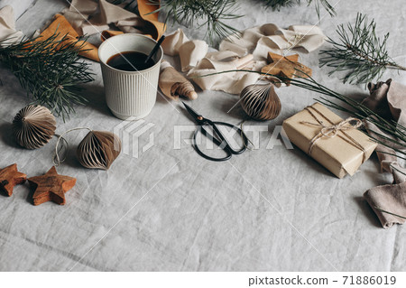 Christmas festive still life composition. Cup of coffee, paper Christmas ornaments and gift box. Velvet ribbons, pine tree branches with scissors. Linen table cloth background. Moody winter banner. Christmas festive still life composition. Cup of coffee, paper Christmas ornaments and gift box. Velvet ribbons, pine tree branches with scissors. Linen table cloth background. Moody winter banner. 71886019