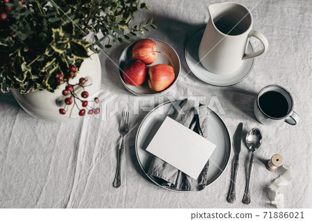 Festive Christmas table setting. Silver cutlery, bowl of apples on linen tablecloth. Cup of coffee, pitcher. Defocused bouquet of holly berries, rose hips. Blank greeting card mockup. Winter design. Festive Christmas table setting. Silver cutlery, bowl of apples on linen tablecloth. Cup of coffee, pitcher. Defocused bouquet of holly berries, rose hips. Blank greeting card mockup. Winter design. 71886021
