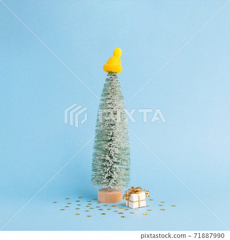 Christmas snowy tree in winter cap and gift on pastel blue background. Minimal New year concept. Magic pine tree Christmas snowy tree in winter cap and gift on pastel blue background. Minimal New year concept. Magic pine tree 71887990