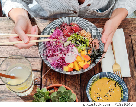 Woman eating tasty colorful healthy natural organic vegetarian Hawaiian poke bowl using asian chopsticks on rustic wooden table. Healthy natural organic eating concept Woman eating tasty colorful healthy natural organic vegetarian Hawaiian poke bowl using asian chopsticks on rustic wooden table. Healthy natural organic eating concept 71889149