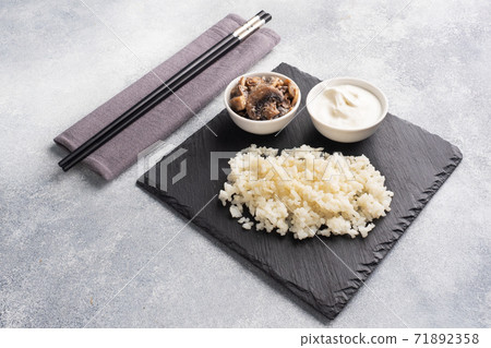 Boiled white rice and mushrooms in sour cream on a black stand with chopsticks. Grey concrete background Copy space. Boiled white rice and mushrooms in sour cream on a black stand with chopsticks. Grey concrete background Copy space. 71892358