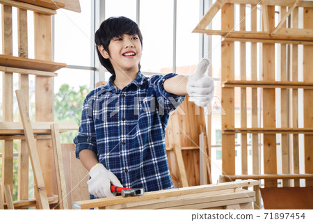 Asian boy with safety glove giving a thumb up and holding hammer in carpentry workshop. Asian boy with safety glove giving a thumb up and holding hammer in carpentry workshop. 71897454