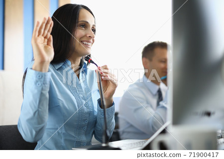 Businesswoman sits at computer with microphone smiling and raising her hand in a welcoming manner. 71900417