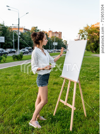 woman artist city summer, background car road, green lawn, begins paint picture on white canvas, hand brush color palette of paints, easel. Clothes white shirt shorts. Hair bun. Side view. 71905304