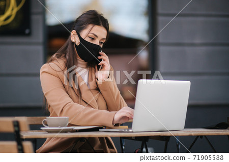Young female in black face mask sitting on terrace in cafe during quarantine. Business woman working outdoos in quarantine. Woman speak by the phone. Covid-19 71905588