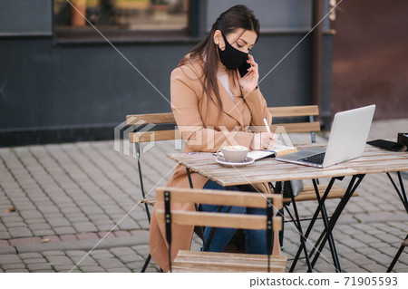 Young female in black face mask sitting on terrace in cafe during quarantine. Business woman working outdoos in quarantine. Woman speak by the phone. Covid-19 Young female in black face mask sitting on terrace in cafe during quarantine. Business woman working outdoos in quarantine. Woman speak by the phone. Covid-19 71905593