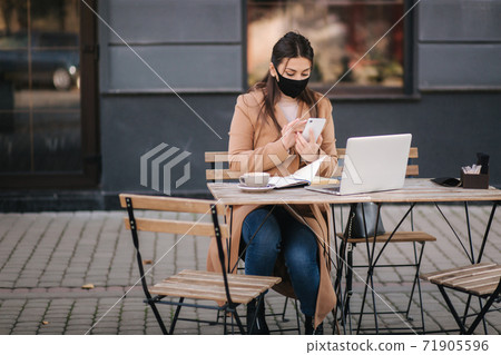 Young female in black face mask sitting on terrace in cafe during quarantine. Business woman working outdoos in quarantine. Woman speak by the phone. Covid-19 Young female in black face mask sitting on terrace in cafe during quarantine. Business woman working outdoos in quarantine. Woman speak by the phone. Covid-19 71905596