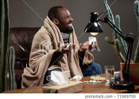 Young man wrapped in a plaid looks sick, ill, sneezing and coughing sitting at home indoors. 71909700