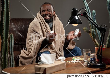 Young man wrapped in a plaid looks sick, ill, sneezing and coughing sitting at home indoors. 71909703