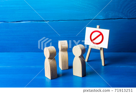 A group of people figurines looking at an easel with a red prohibitory symbol NO. Expression of protest and disagreement. Inaccessibility and absence. Quarantine and lockdown. Hard restrictions A group of people figurines looking at an easel with a red prohibitory symbol NO. Expression of protest and disagreement. Inaccessibility and absence. Quarantine and lockdown. Hard restrictions 71913251