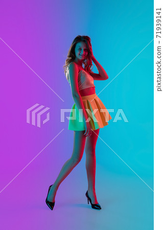 Beautiful seductive girl in fashionable bright summer outfit posing on colour gradient pink-blue background in neon light. Beautiful seductive girl in fashionable bright summer outfit posing on colour gradient pink-blue background in neon light. 71939141