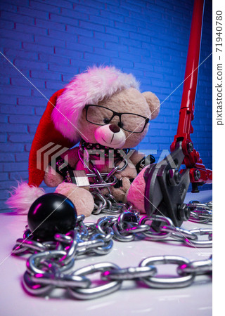 a toy bear wearing leather belts and a Santa Claus hat in the New year Christmas is chained and handcuffed an accessory for BDSM games next to a bolt cutter 71940780