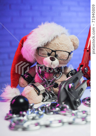 a toy bear wearing leather belts and a Santa Claus hat in the New year Christmas is chained and handcuffed an accessory for BDSM games next to a bolt cutter 71940809