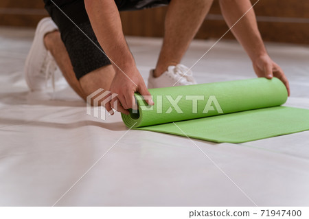 Strong handsome young man, an athlete, unwinds a roll mat, preparing a place for sports. Determined guy doing physical training 71947400