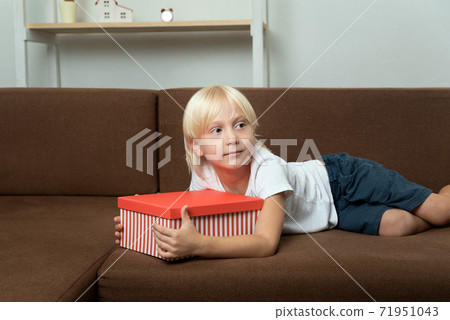 Child lies on the couch with gift box in his hands. Blond boy holding bright box in his hands 71951043