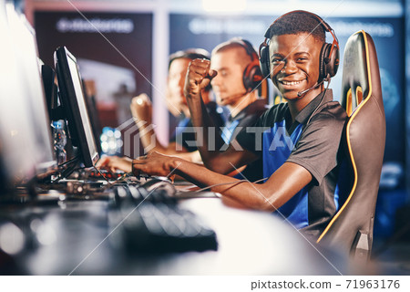 Side view of a cheerful african guy, professional cybersport gamer wearing headphones looking at camera and smiling while participating with team in eSport tournament 71963176