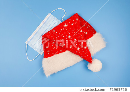 Santa Claus red hat and blue protective medical mask on blue table. Top view. Santa Claus red hat and blue protective medical mask on blue table. Top view. 71965736