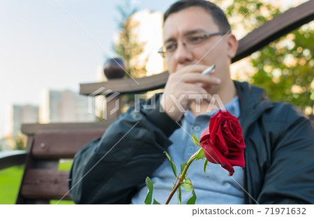 red rosebud in hand and against the background of a thoughtful guy who is waiting for a girl on a date and smoking a cigarette red rosebud in hand and against the background of a thoughtful guy who is waiting for a girl on a date and smoking a cigarette 71971632