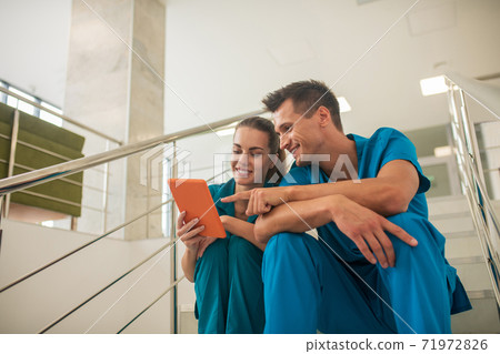 Two medical workers sitting on the stairs and looking at something on tablet Two medical workers sitting on the stairs and looking at something on tablet 71972826