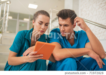 Two medical workers sitting on the stairs and looking at something on tablet Two medical workers sitting on the stairs and looking at something on tablet 71972828