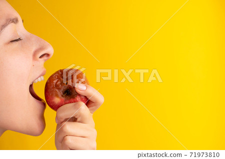 The girl bites a rotten apple with a worm on a yellow background. Expired products, junk food The girl bites a rotten apple with a worm on a yellow background. Expired products, junk food 71973810