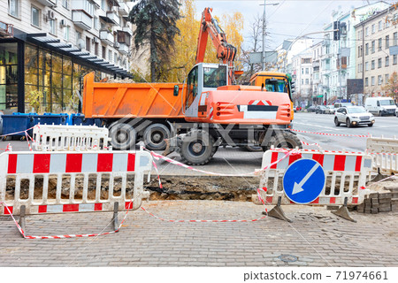 Heavy road machinery, an excavator and a truck on a fenced section of a city street sidewalk are involved in the repair of a heating main. Heavy road machinery, an excavator and a truck on a fenced section of a city street sidewalk are involved in the repair of a heating main. 71974661