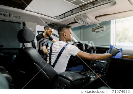 Car service and detailing station. Back angle indoor shot of two professional multiethnic men workers, providing a vehicle interior cleaning, wiping front panel with microfiber cloth and brush Car service and detailing station. Back angle indoor shot of two professional multiethnic men workers, providing a vehicle interior cleaning, wiping front panel with microfiber cloth and brush 71974923