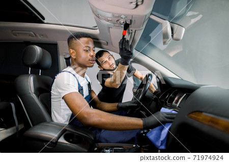 Professional car detailing and maintenance concept. Two multiethnica young male workers cleaning vehicle interior with special cloth and brush at the car service station. Selective focus Professional car detailing and maintenance concept. Two multiethnica young male workers cleaning vehicle interior with special cloth and brush at the car service station. Selective focus 71974924