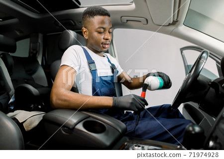 Portrait of young African male car detailing worker, sitting inside of modern vehicle, pouring plastic trim care milk on the soft brush. Car detailing, plastic care Portrait of young African male car detailing worker, sitting inside of modern vehicle, pouring plastic trim care milk on the soft brush. Car detailing, plastic care 71974928