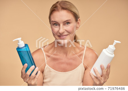Portrait of a beautiful mature woman choosing between two cosmetic products while standing isolated over beige background in studio 71976486