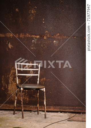 A rusty chair with peeling paint and a shabby leatherette seat against a rusty wall on the concrete floor. 71977947