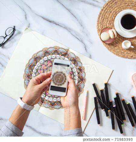 Woman is taking photos of colored mandala from sketchbook on white marble desk with displayed object Woman is taking photos of colored mandala from sketchbook on white marble desk with displayed object 71983393