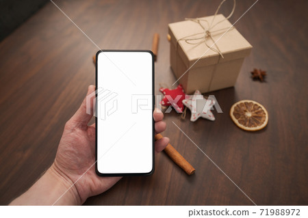 Man hand hold smarthone with white screen with christmas decorations on background Man hand hold smarthone with white screen with christmas decorations on background 71988972