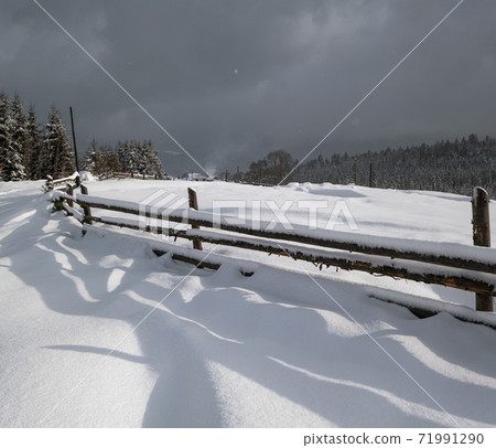 Picturesque waved shadows on snow from wood fence. Alpine mountain winter hamlet outskirts, snowy path, fir forest on far misty and cloudy hills. Picturesque waved shadows on snow from wood fence. Alpine mountain winter hamlet outskirts, snowy path, fir forest on far misty and cloudy hills. 71991290