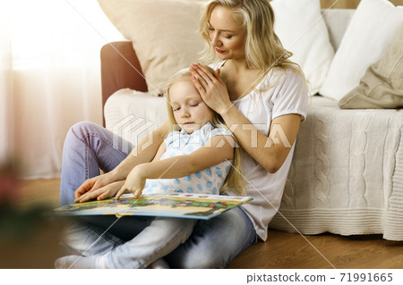 Happy family. Blonde young mother reading a book to her cute daughter while sitting at wooden floor in sunny room. Motherhood concept Happy family. Blonde young mother reading a book to her cute daughter while sitting at wooden floor in sunny room. Motherhood concept 71991665