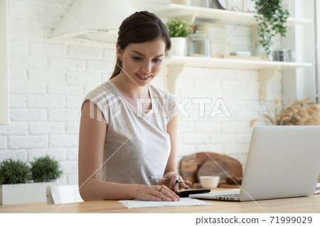 Smiling young woman checking finances, using laptop, sitting in kitchen 71999029