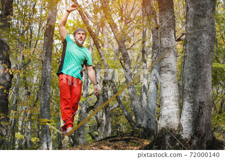 A bearded man in age balances while sitting on a taut slackline in the autumn forest. Outdoor Leisure 72000140