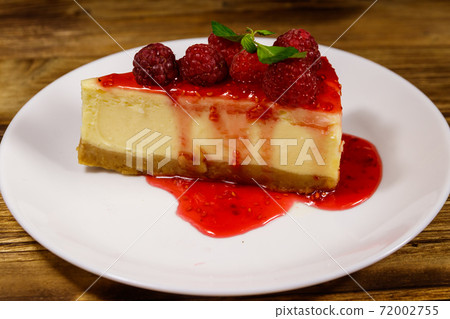 Piece of tasty New York cheesecake with raspberries and raspberry jam in a white plate on wooden table Piece of tasty New York cheesecake with raspberries and raspberry jam in a white plate on wooden table 72002755