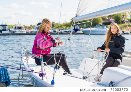 Two young female athletes prepare a sailing boat for the regatta on the river pier 72003770