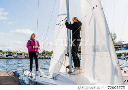 Two young female athletes prepare a sailing boat for the regatta on the river pier Two young female athletes prepare a sailing boat for the regatta on the river pier 72003775