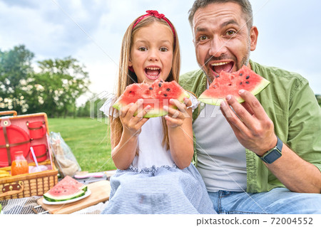 Portrait of happy dad and his adorable little daughter looking at camera while eating watermelon, family having a picnic in the green park on a summer day Portrait of happy dad and his adorable little daughter looking at camera while eating watermelon, family having a picnic in the green park on a summer day 72004552