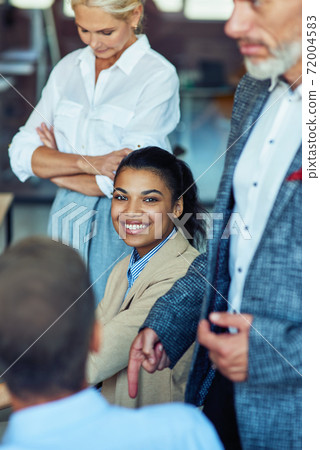 Young cheerful mixed race woman, female office worker smiling at camera while having a meeting with colleagues Young cheerful mixed race woman, female office worker smiling at camera while having a meeting with colleagues 72004583