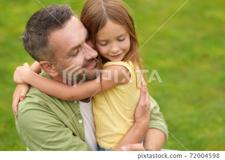 Portrait of happy young father and adorable little daughter hugging each other, standing with eyes closed while spending time in the green park on a summer day Portrait of happy young father and adorable little daughter hugging each other, standing with eyes closed while spending time in the green park on a summer day 72004598