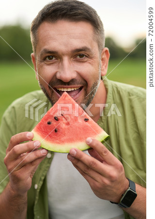 Happy young man looking at camera while eating watermelon in the green park on a summer day Happy young man looking at camera while eating watermelon in the green park on a summer day 72004599
