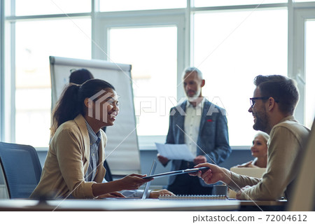 Two cheerful diverse office workers discussing something and smiling while having a meeting with colleagues in the modern office Two cheerful diverse office workers discussing something and smiling while having a meeting with colleagues in the modern office 72004612