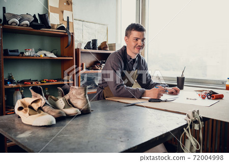 A young shoemaker makes a drawing for a pattern for leather shoes on a table in his workshop 72004998