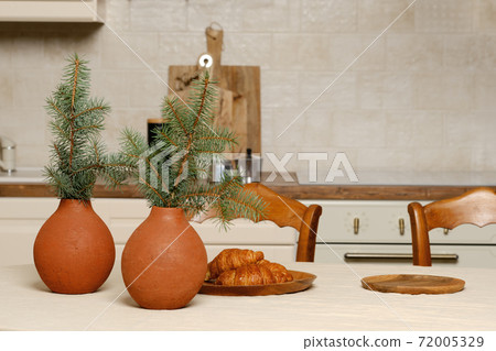 Kitchen table with croissants on plate and clay vases with fir branches Kitchen table with croissants on plate and clay vases with fir branches 72005329