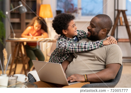 Happy affectionate African boy embracing his father with laptop against daughter Happy affectionate African boy embracing his father with laptop against daughter 72005777