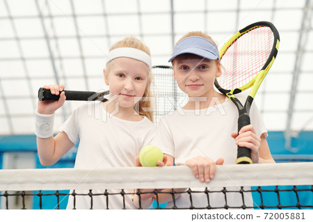 Two cute friendly girls in white sportswear holding tennis rackets on shoulders Two cute friendly girls in white sportswear holding tennis rackets on shoulders 72005881
